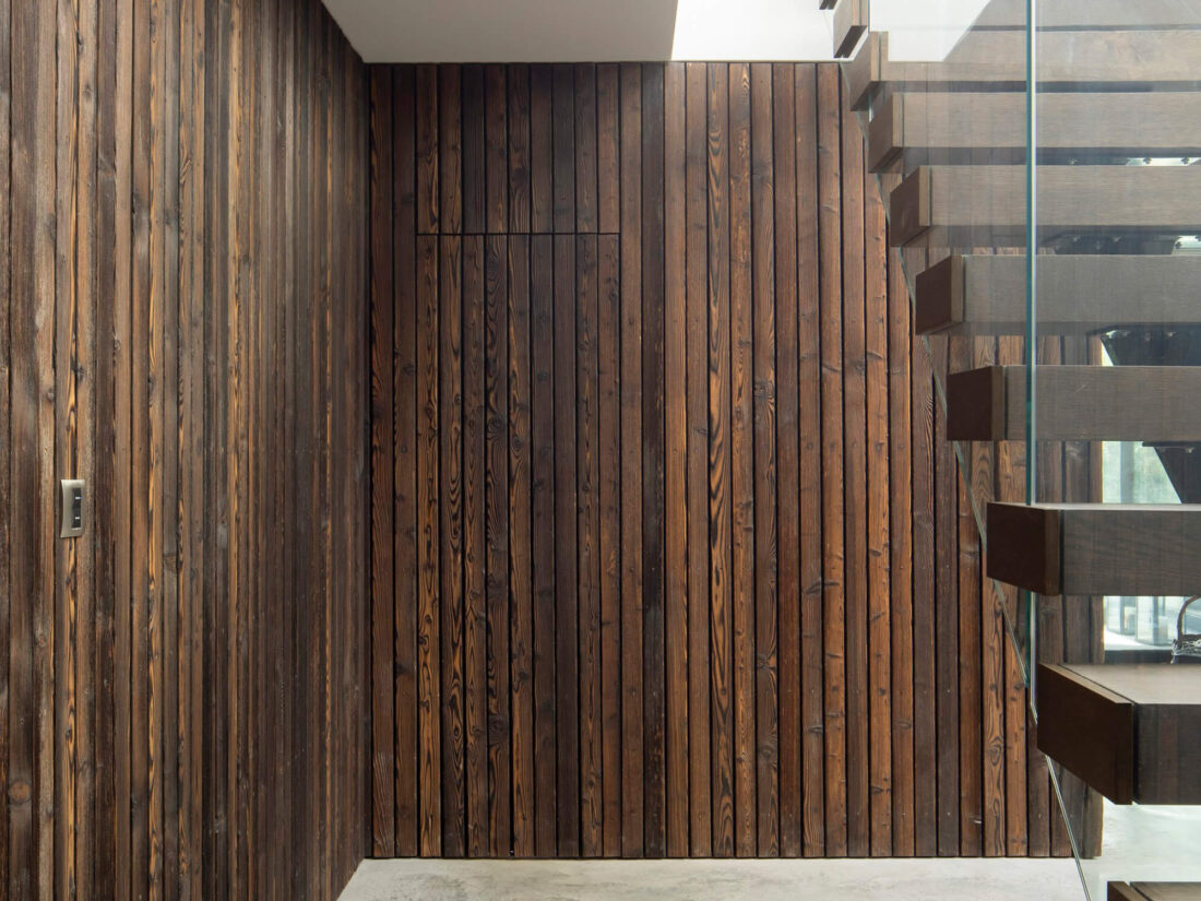 Interior view of a modern space featuring vertical Degmeda "Amber" larch charred wood cladding on the walls, including a hidden door, paired with a dark wood and glass staircase.
