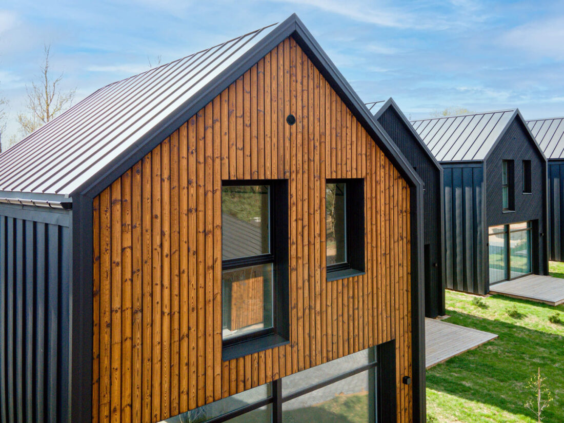 Exterior view of modern gabled residences at "Ežero vilos" in Lithuania featuring vertical Degmeda "Amber" spruce charred wood cladding, contrasted with black standing seam metal roofing and side facades.