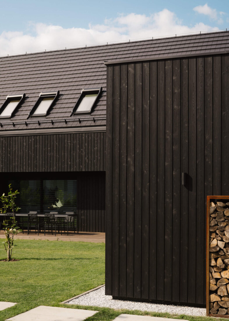 Architectural detail of a modern residence in Tuszynek featuring vertical Degmeda "Dark Black" larch charred wood cladding. The brushed matte texture of the Shou Sugi Ban boards is shown alongside a corten steel wood storage unit and a dark tiled roof with skylights.