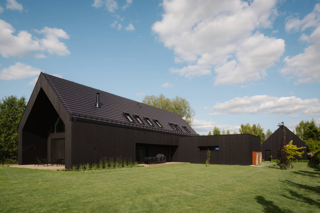 Exterior view of a modern gabled residence in Tuszynek featuring vertical Degmeda "Dark Black" larch charred wood cladding. The matte black finish of the Shou Sugi Ban boards is paired with a matching dark tiled roof and a corten steel wood storage accent, set against an expansive green lawn.