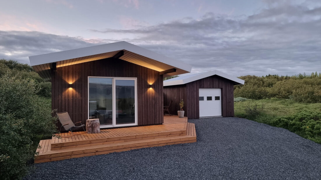Modernių poilsio namelių eksterjeras su C klasės Degmeda "Dark Brown" deginto maumedžio dailylentėmis, pabrėžiant tamsaus fasado ir šviesios medinės terasos kontrastą vakaro apšvietime.