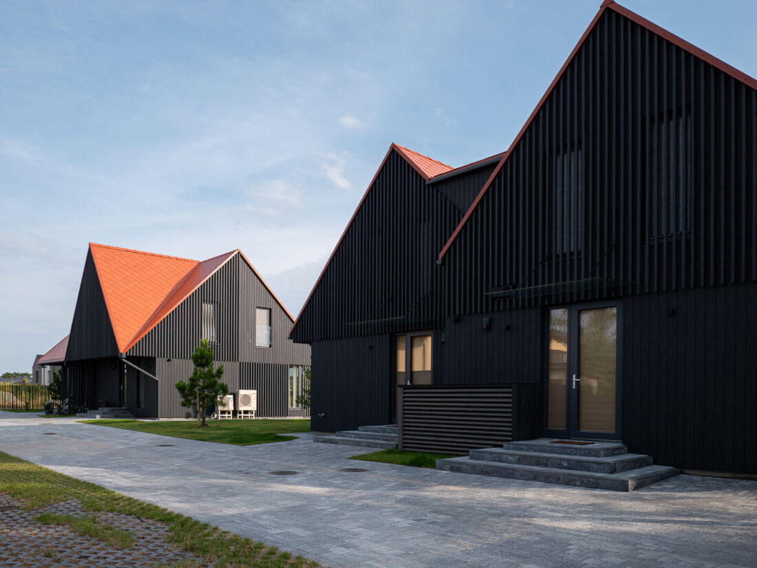 Exterior view of modern gabled residential houses in Palanga featuring vertical Degmeda "Dark Grey" spruce charred wood cladding. The deep grey Shou Sugi Ban boards create a sharp material contrast with the vibrant orange clay tile roofs and light stone paving.