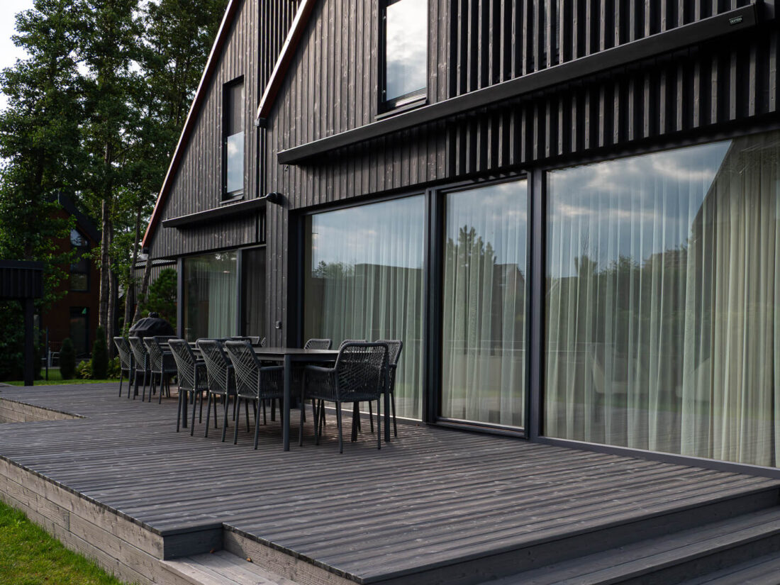View of an outdoor terrace and residential facade featuring Degmeda "Dark Grey" spruce charred wood boards. The vertical cladding and horizontal terrace decking display a brushed grain texture, contrasting with large glass sliding doors and modern dark grey patio furniture.