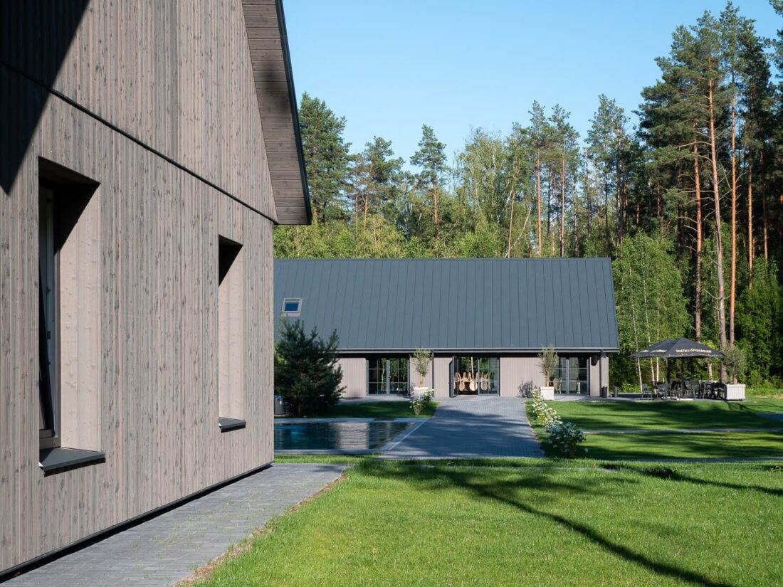 Perspective view of the "Parko vilos" resort architecture in Panemunė featuring vertical Degmeda "Light Grey" spruce charred wood cladding on modern gabled buildings, integrated into a natural forest setting with a pool and paved pathways.