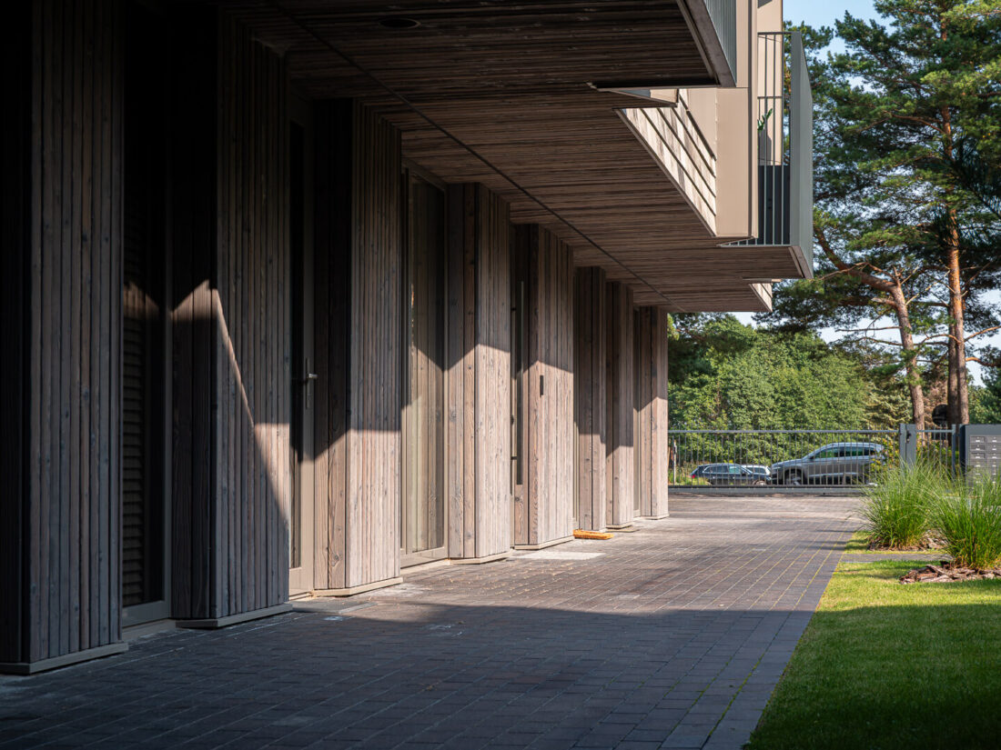 Ground-level architectural detail of a residential building in Palanga featuring Degmeda "Light Grey" spruce charred wood cladding. The brushed Shou Sugi Ban boards are applied vertically to facade pillars and horizontally to balcony soffits, creating a textured contrast with the dark stone paving and glass entrances.