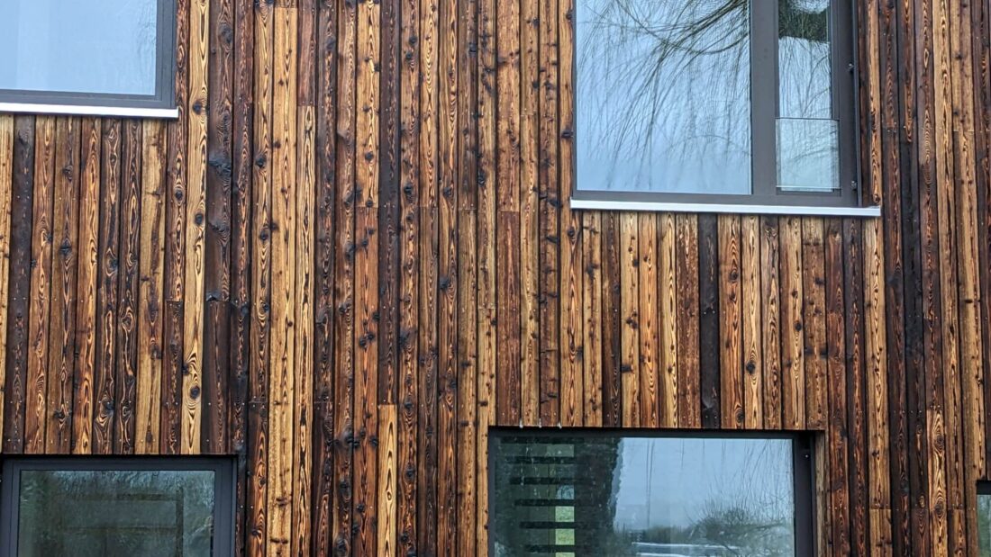 Detailed view of a building facade featuring vertical C-class Degmeda "Natural" brushed charred larch wood cladding, highlighting the intense textured grain and material contrast with large glass windows.
