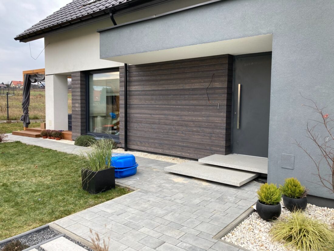 Exterior view of a modern home entrance showcasing horizontal C-class Degmeda "Natural" charred wood cladding installed alongside grey plastered facade elements and a dark grey door.