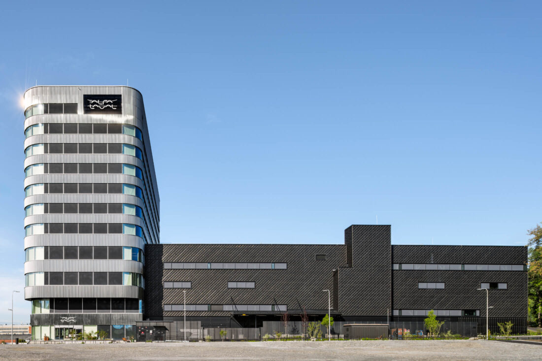Wide exterior view of the Alfa Laval office and innovation center (Separatorn 1) in Stockholm featuring Degmeda "Deep Char" spruce charred wood cladding. The dark, cracked carbon texture of the Yakisugi boards is installed in a diamond-grid pattern on the facade, contrasting with the building's curved metallic sections and large glass windows.