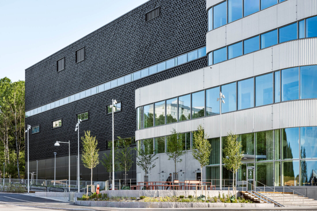 Detailed view of a commercial building facade in Stockholm featuring Degmeda "Deep Char" spruce charred wood cladding. The crackled, matte black texture of the Yakisugi boards is installed in a diagonal grid pattern, providing a material contrast to the adjacent silver metallic corrugated panels and large glass windows.