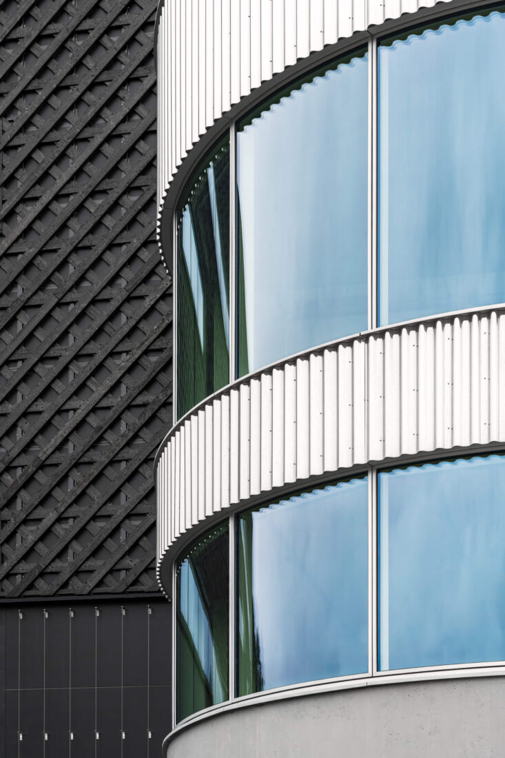Architectural detail of a commercial building facade showing Degmeda "Deep Char" spruce charred wood cladding in a diagonal grid pattern. The heavily crackled matte texture of the Shou Sugi Ban boards contrasts with a curved section of white corrugated metal panels and reflective glass windows.