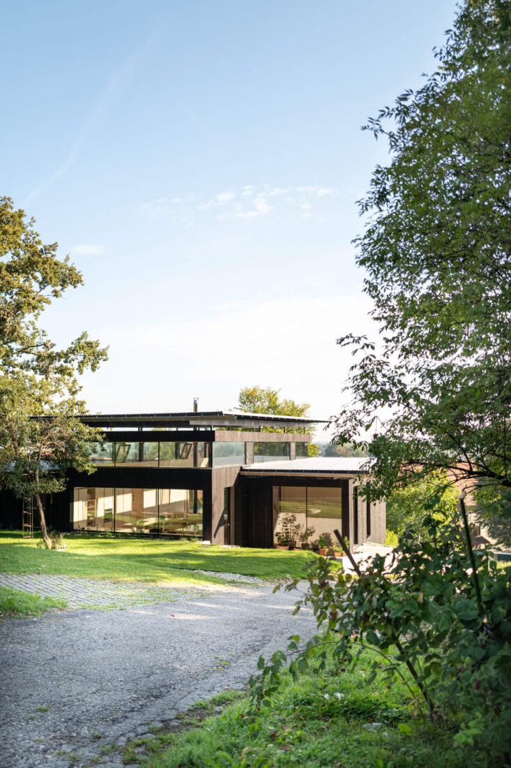 Exterior side view of the House Above the City in Slovenia featuring vertical Degmeda "Dark Black" larch charred wood boards and wide panoramic windows reflecting the green hillside landscape.