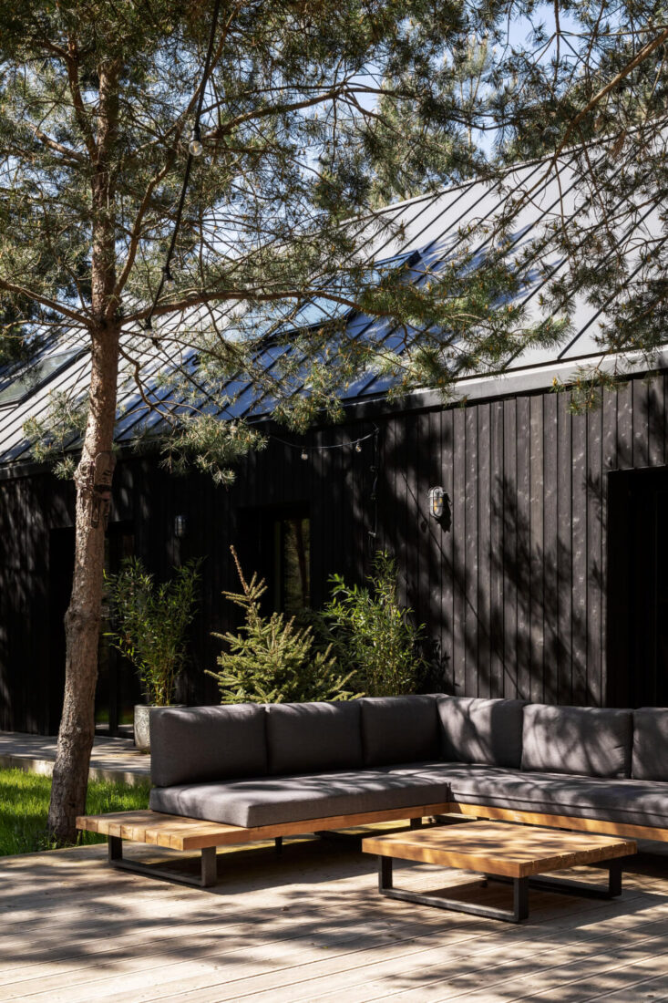Exterior view of a residential family home in Poland featuring vertical Degmeda Dark Black larch charred wood cladding and a wooden terrace with outdoor furniture set in a forest landscape.