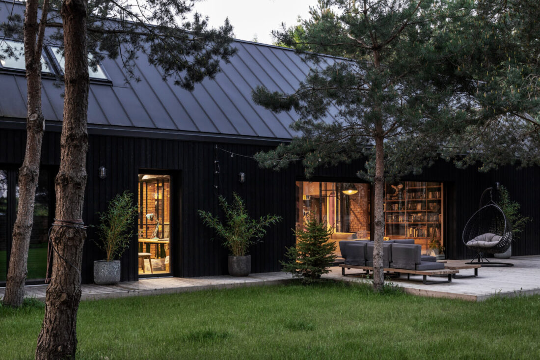 Exterior dusk view of a forest family home in Poland showing Degmeda "Dark Black" larch charred wood cladding on the facade and a wooden terrace with outdoor furniture under a steep metal roof.
