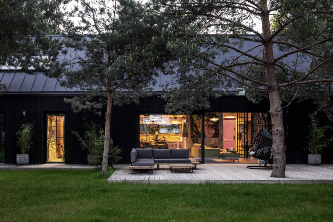 Exterior view of a modern forest residence in Poland at dusk featuring vertical Degmeda "Dark Black" larch charred wood cladding and an illuminated wooden terrace with outdoor furniture.