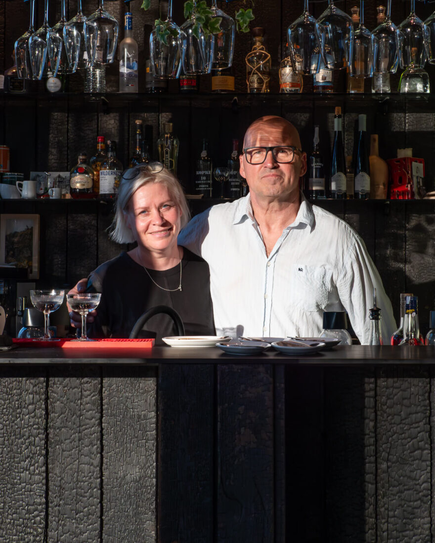 The owners of Velo Bar in Lithuania standing behind a bar counter and back wall clad in vertical Degmeda "Deep Char" larch charred wood boards.