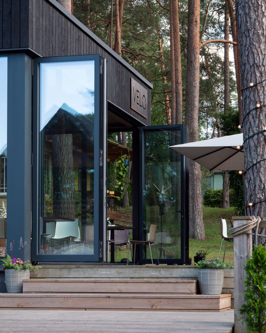 Entrance of Velo Bar in Lithuania featuring vertical Degmeda "Deep Char" larch cladding, folding glass doors, and a timber terrace surrounded by forest.