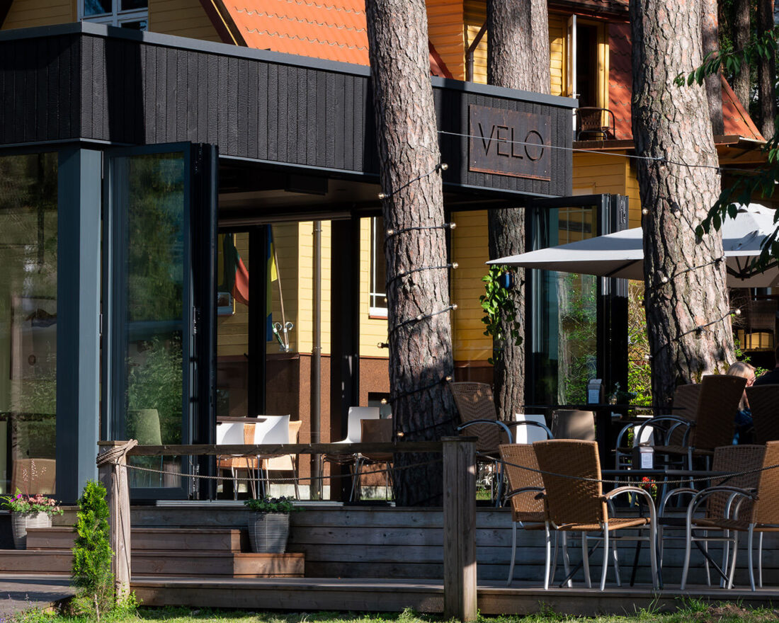 Exterior view of the Velo Bar pavilion in Kačerginė, Lithuania, featuring vertical Degmeda "Deep Char" larch charred wood cladding integrated with large folding glass doors and situated within a pine forest landscape.