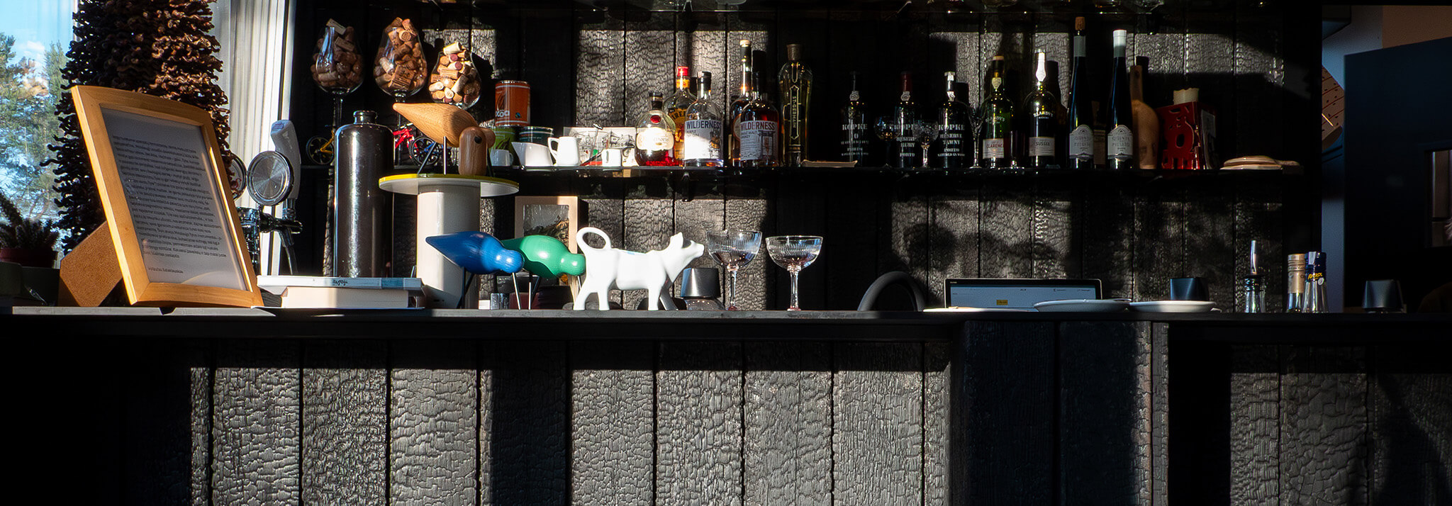 Interior panorama of Velo Bar showing a long bar counter and back shelving unit clad in Degmeda "Deep Char" larch charred wood boards with side lighting highlighting the heavy texture.