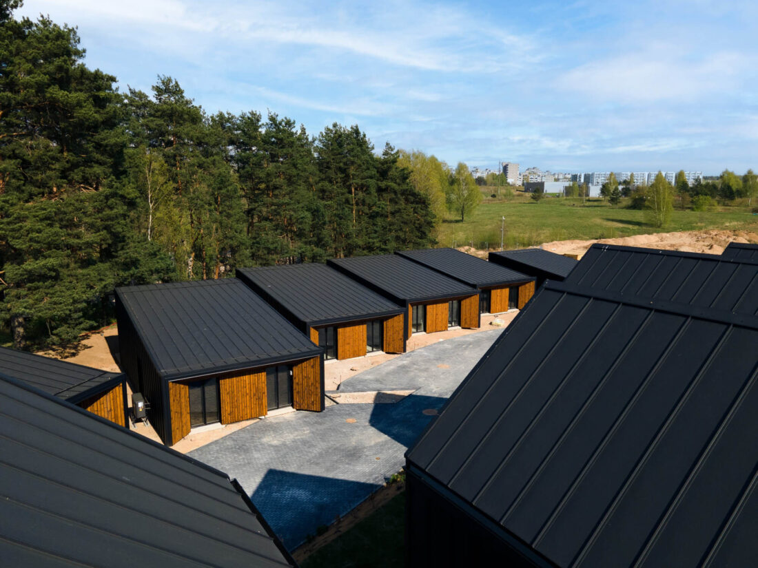 High-angle aerial view of gabled houses in the Lake Villas development, Vilnius, featuring Degmeda "Amber" spruce boards and black standing seam metal roofs bordered by a pine forest.