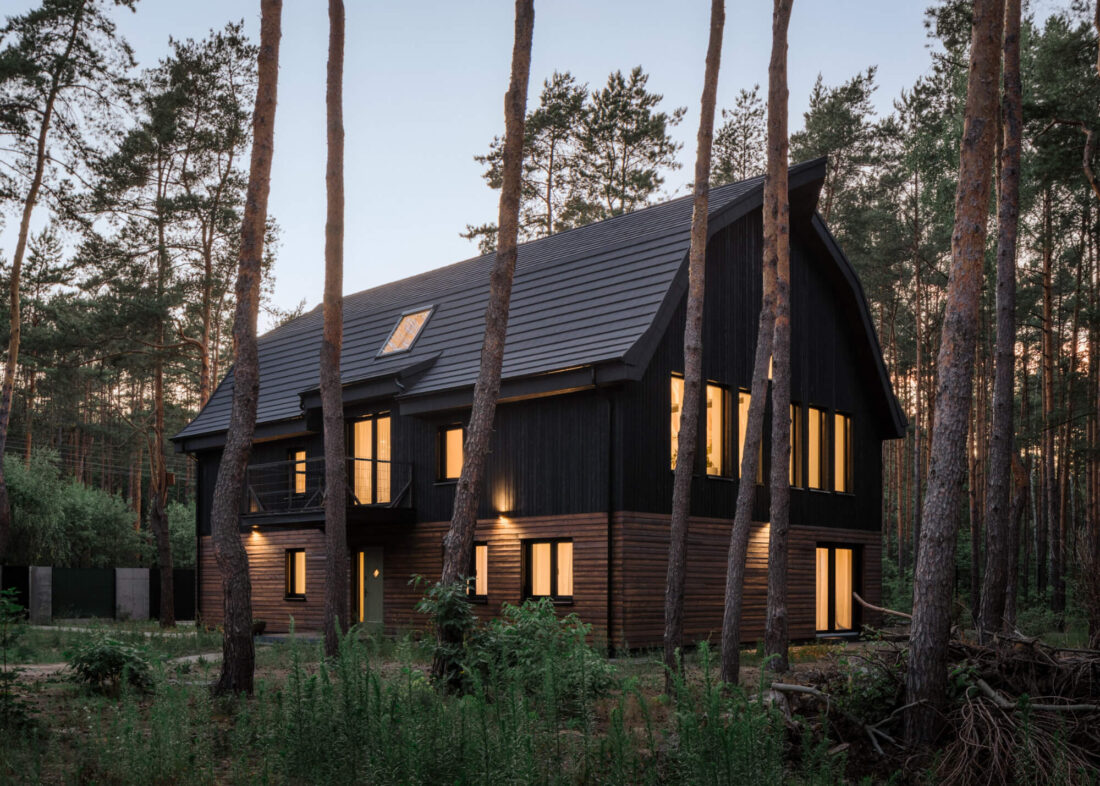 Dusk view of the Wawer Barn in Poland nestled among pine trees featuring vertical Degmeda Dark Black larch boards on the upper level and horizontal Amber larch boards on the ground floor with lit windows.