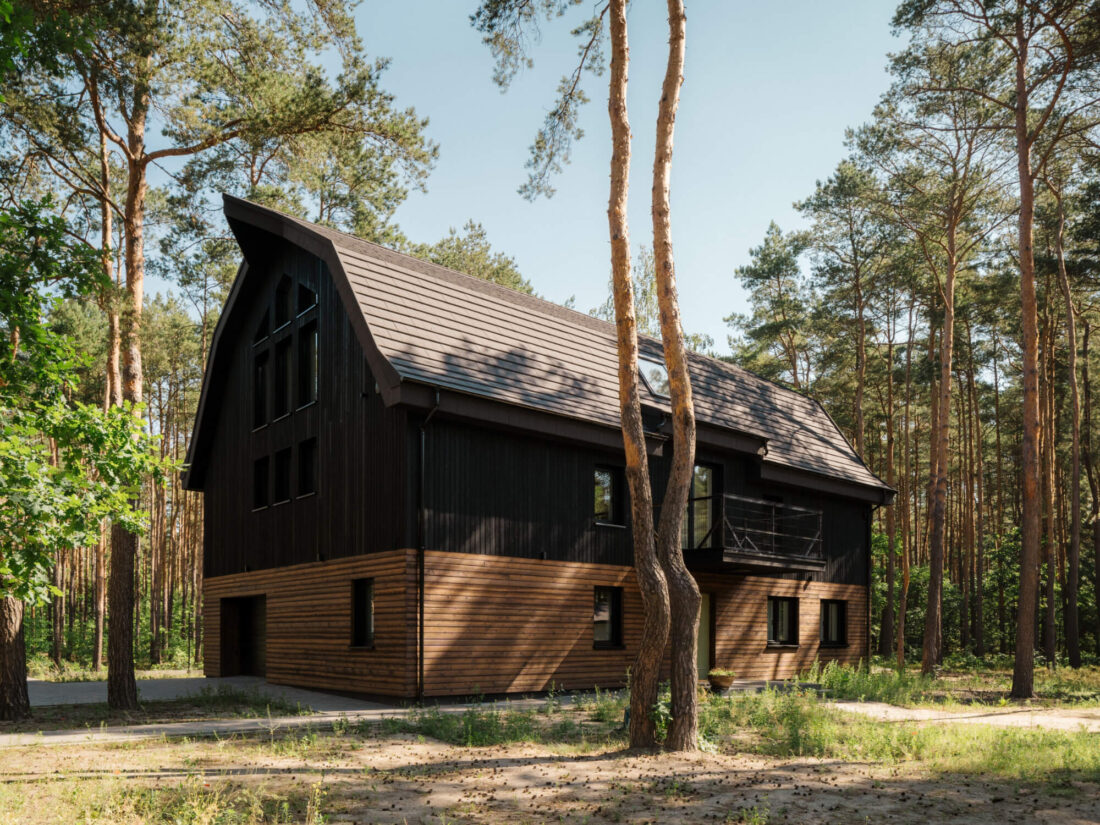 Exterior view of the Wawer Barn residence in Poland featuring vertical Degmeda "Dark Black" larch boards on the upper level and horizontal "Amber" larch boards at the base, set within a pine forest.