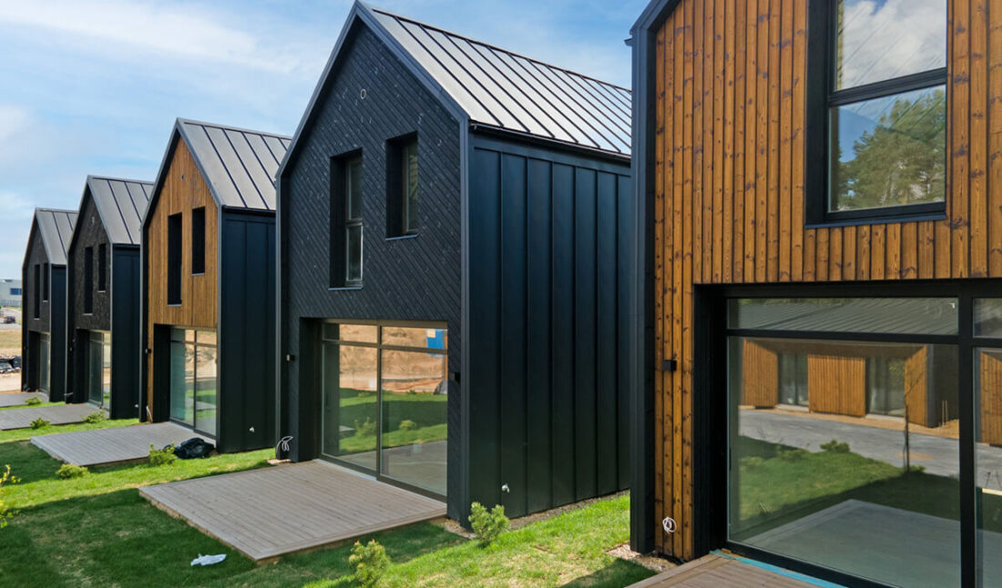 Perspective view of the Lake Villas development in Vilnius featuring a row of gabled houses clad in vertical Degmeda "Dark Black" and "Amber" spruce boards with standing seam metal roofs.