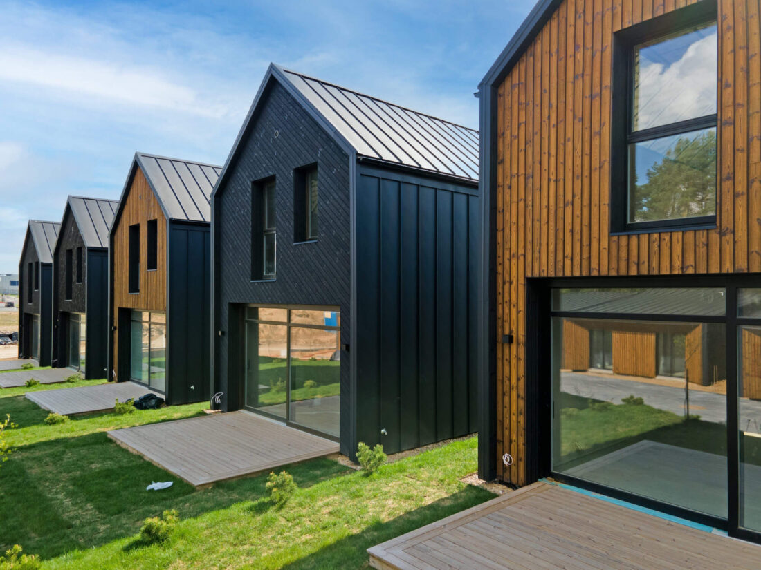Perspective view of modern gabled houses at the Lake Villas development in Vilnius featuring the aesthetic contrast of vertical Degmeda "Dark Black" and "Amber" spruce boards and wooden terraces.