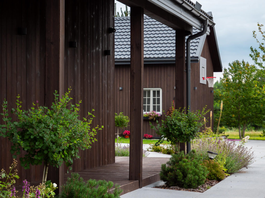 Exterior view of a residential building in Elektrėnai featuring vertical Degmeda Dark Brown larch boards on the facade and porch columns integrated with garden landscaping.
