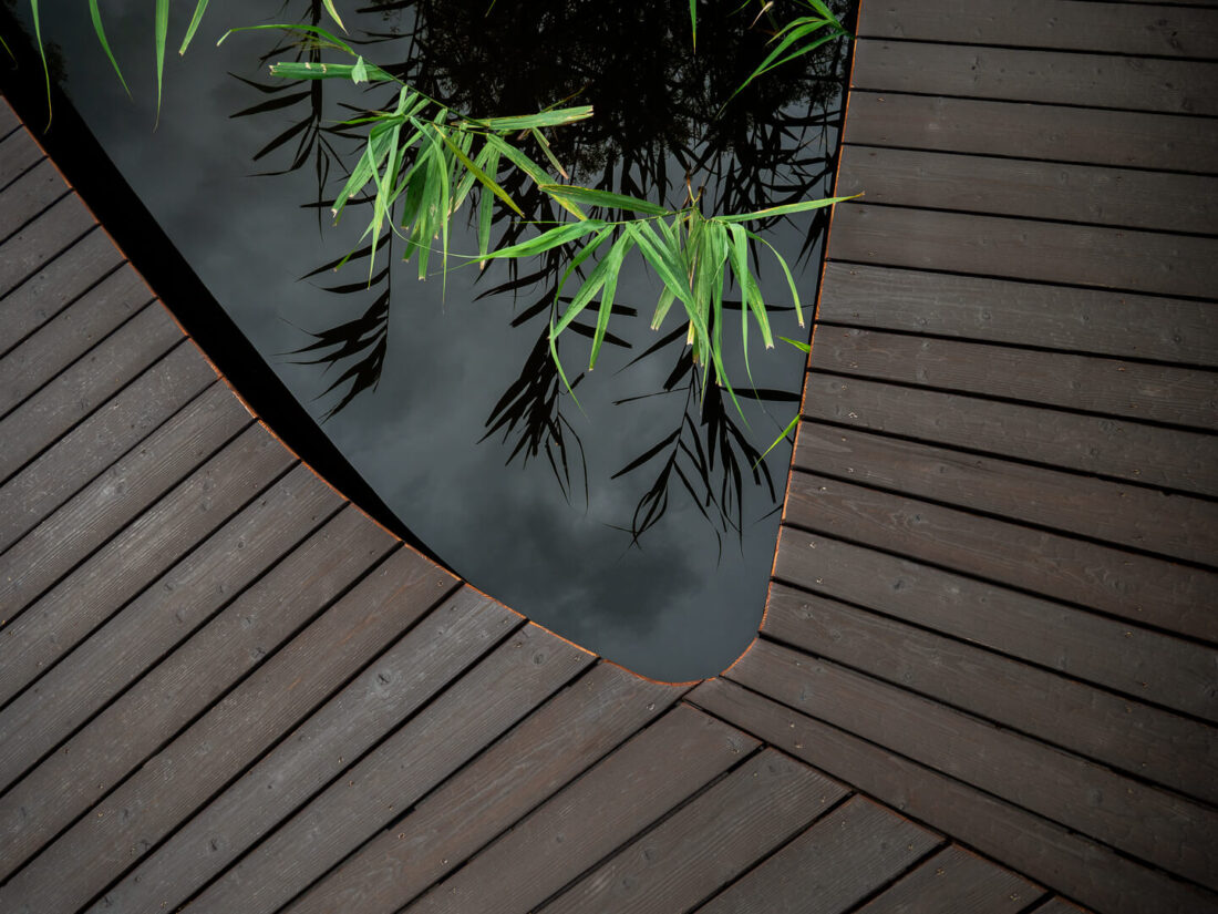 Top-down detail of a lakeside footbridge featuring Degmeda Dark Brown larch boards meeting the water edge with reflections of reeds.