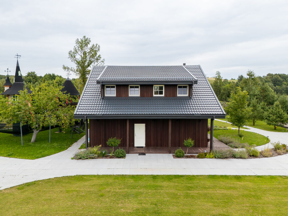 Elevated front elevation of a traditional gabled house in Elektrėnai featuring vertical Degmeda Dark Brown larch boards, three dormer windows, and a black tiled roof.