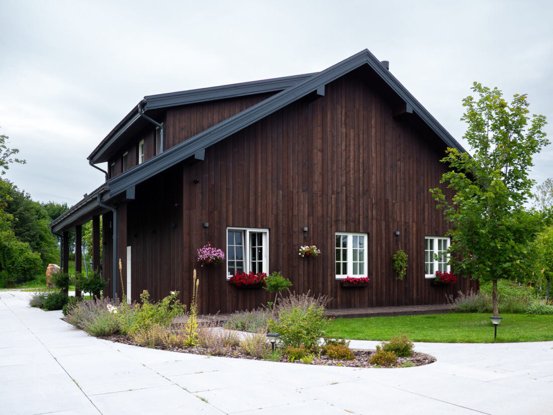 Exterior view of a traditional gabled house in Elektrėnai featuring vertical Degmeda Dark Brown larch boards, white window frames with flower boxes, and garden landscaping.