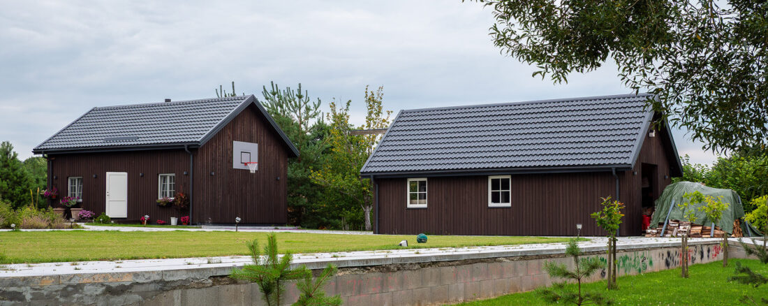 Exterior view of matching gabled buildings at a residential complex in Lithuania featuring vertical Degmeda "Dark Brown" larch boards and grey tile roofs set in a landscaped garden.