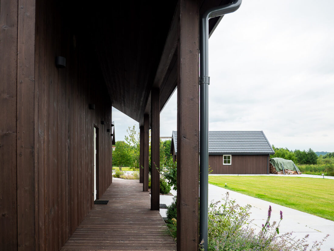 Perspective view of a covered terrace at a residential complex in Elektrėnai featuring vertical Degmeda Dark Brown larch boards and matching timber columns.