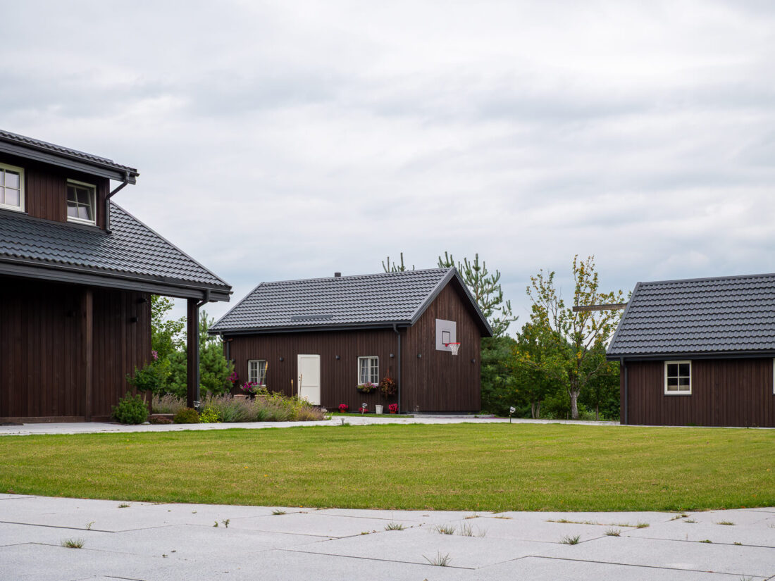 Exterior view of a traditional residential complex in Lithuania featuring multiple buildings clad in vertical Degmeda Dark Brown larch boards with pitched grey tile roofs.