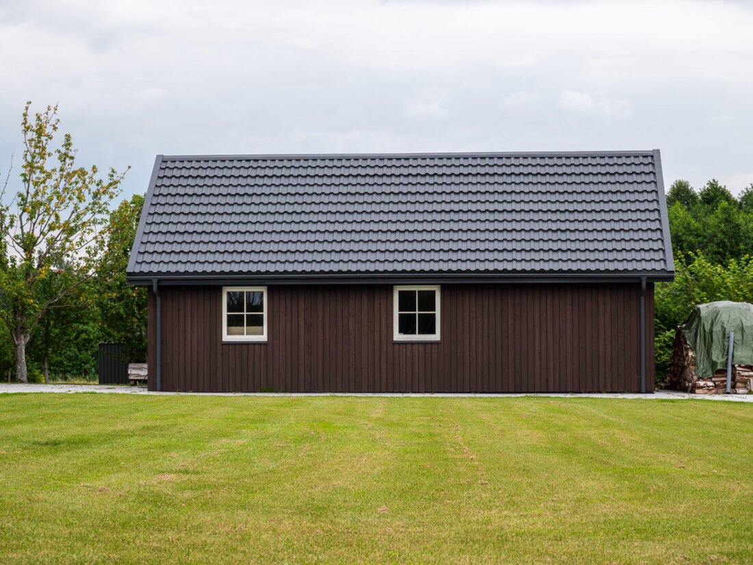Side elevation of a traditional gabled house in Lithuania featuring vertical Degmeda Dark Brown larch boards and a grey tiled roof overlooking a green lawn.
