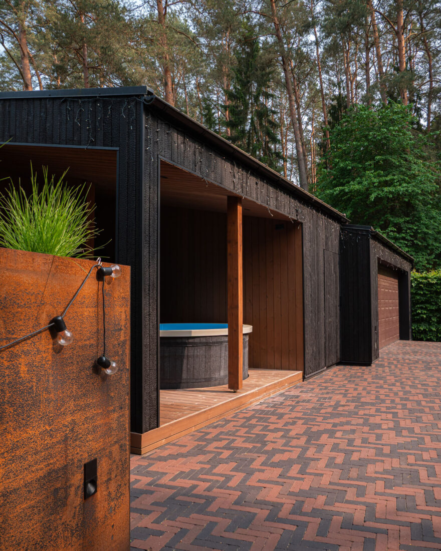 Exterior view of an outbuilding at the Kačerginė residence featuring a hot tub area clad in vertical Degmeda Deep Char and Amber larch charred wood boards with a brick paved driveway.