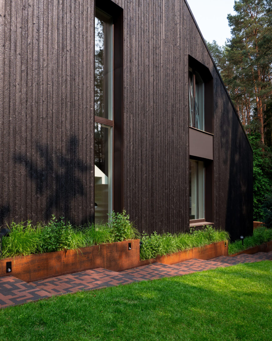 Close-up facade view of the Kačerginė residence featuring vertical Degmeda "Deep Char" larch charred wood cladding positioned above weathered corten steel planters and a brick garden path.