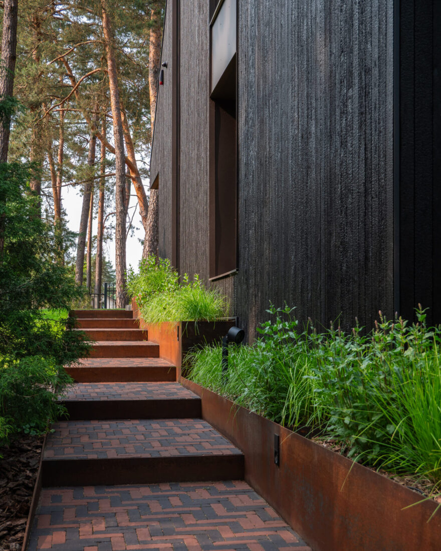 Side view of the Kačerginė residence facade featuring vertical Degmeda "Deep Char" larch charred wood cladding and a garden path with weathered corten steel steps.