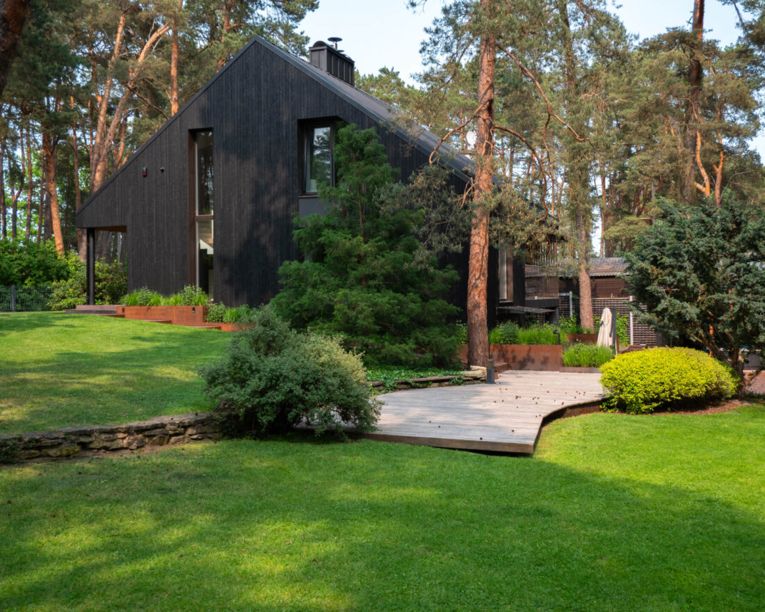 Exterior view of a modern residence in Kačerginė featuring vertical Degmeda "Deep Char" larch charred wood cladding and a curved wooden deck integrated into a landscaped forest garden.