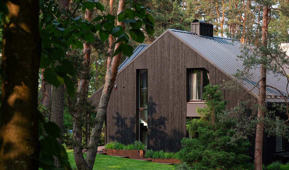 Exterior view of a modern residence in Kačerginė, Lithuania, featuring vertical Degmeda "Deep Char" larch charred wood boards and a dark pitched metal roof nestled among mature pine trees.