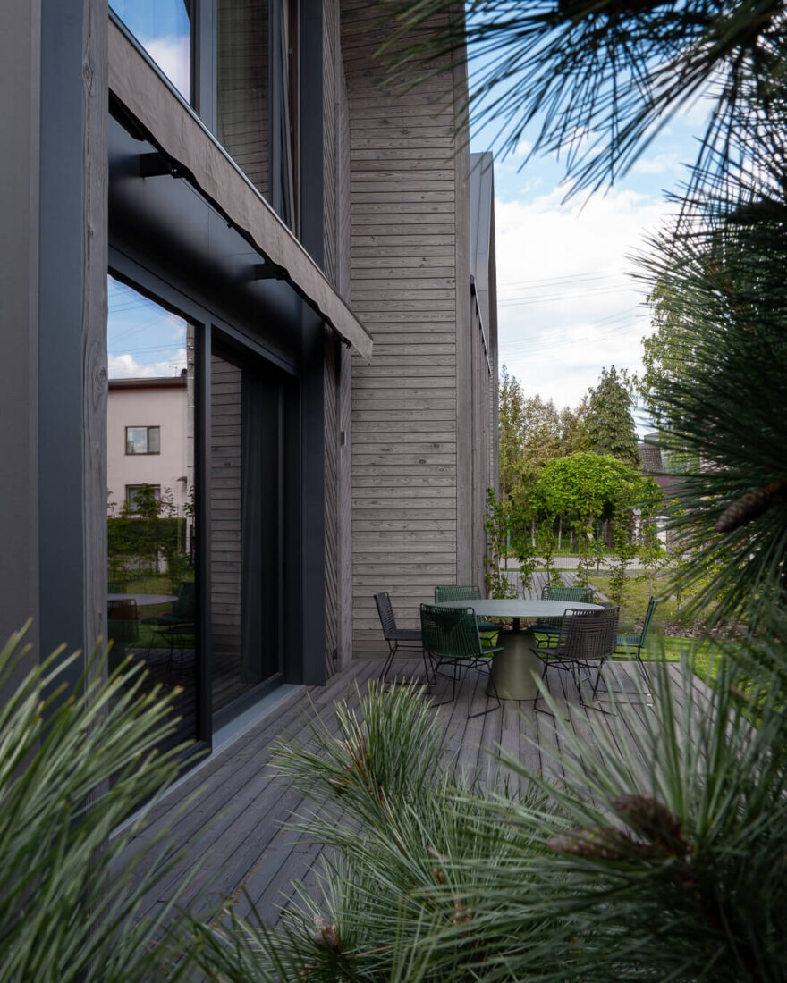 View through pine branches of a modern house facade in Kaunas featuring Degmeda "Light Grey" spruce boards in mixed horizontal and chevron patterns adjacent to an outdoor terrace.