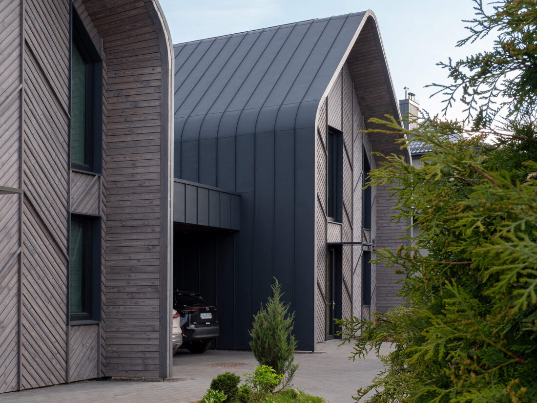 Exterior view of a modern semi-detached house in Kaunas, featuring Degmeda Light Grey spruce facade boards installed in a distinctive chevron pattern, contrasted by a sleek dark grey curved metal roof.
