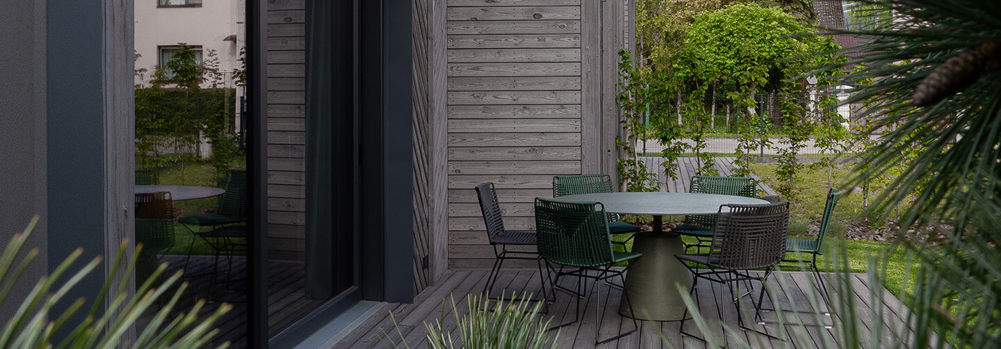 Exterior terrace view of a modern residence in Kaunas featuring Degmeda "Light Grey" spruce facade boards in horizontal and chevron patterns adjacent to a wooden deck with outdoor furniture.