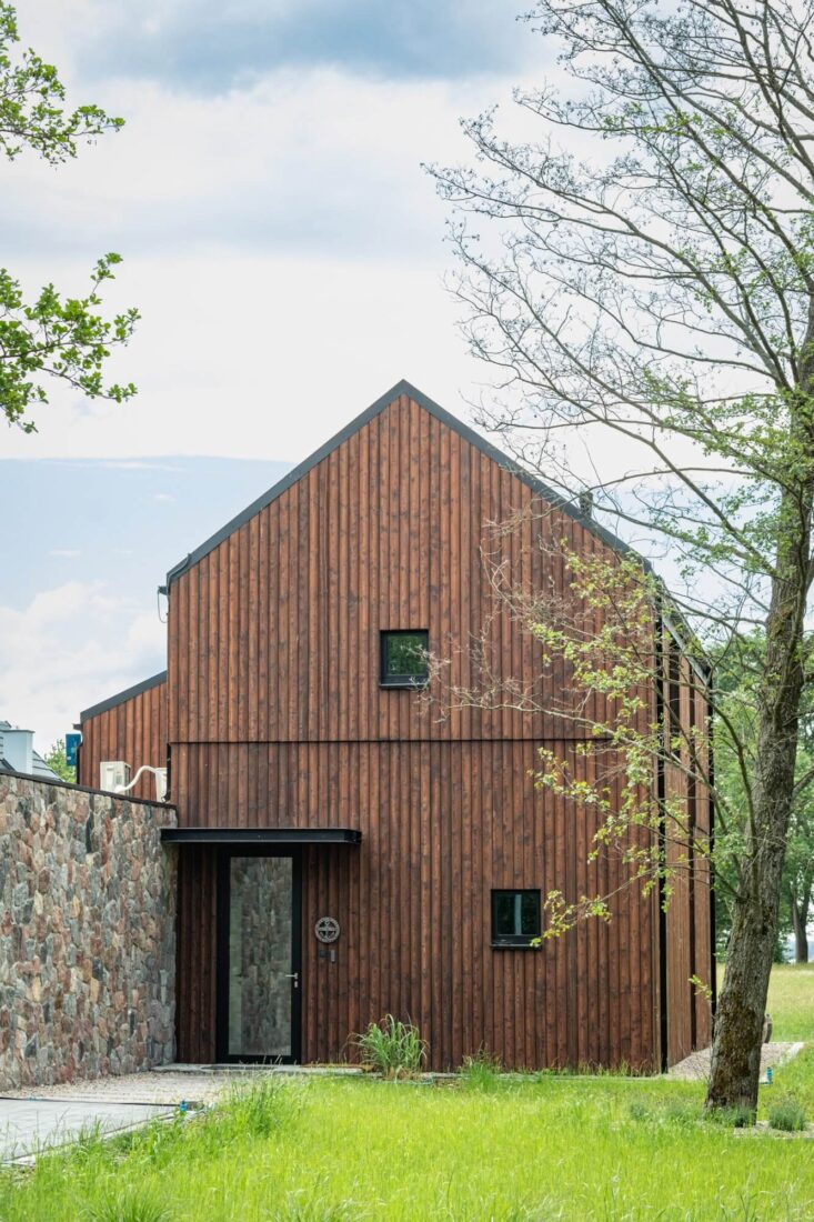 Gabled side facade of the Glass Gable House in Poland clad with vertical Degmeda "Red Amber" larch charred wood boards adjacent to a natural stone wall.