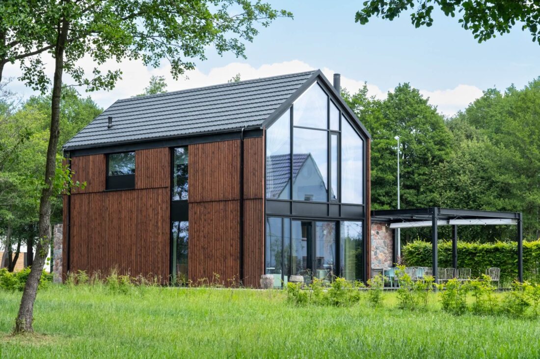Exterior view of the Glass Gable House in Poland featuring vertical Degmeda "Red Amber" larch charred wood boards and a full-height glass gabled facade overlooking a green field.