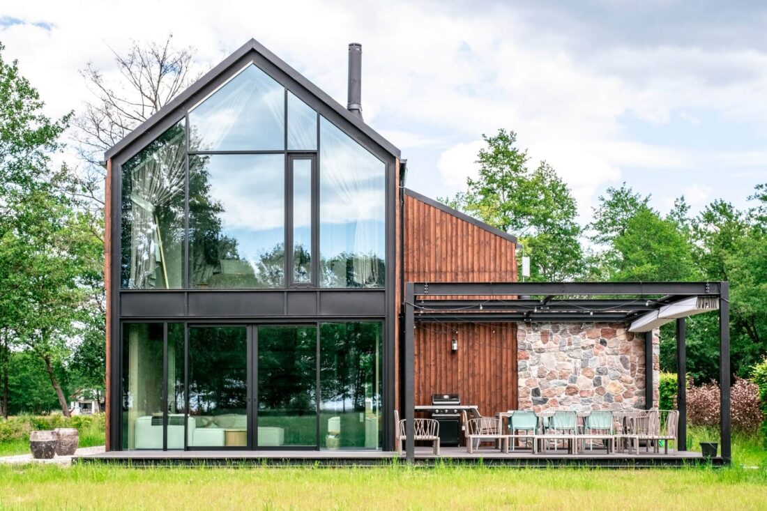 Exterior view of the Glass Gable House in Poland featuring vertical Degmeda "Red Amber" larch charred wood boards, a large glass gable facade, and an outdoor terrace with a stone wall and dining area.